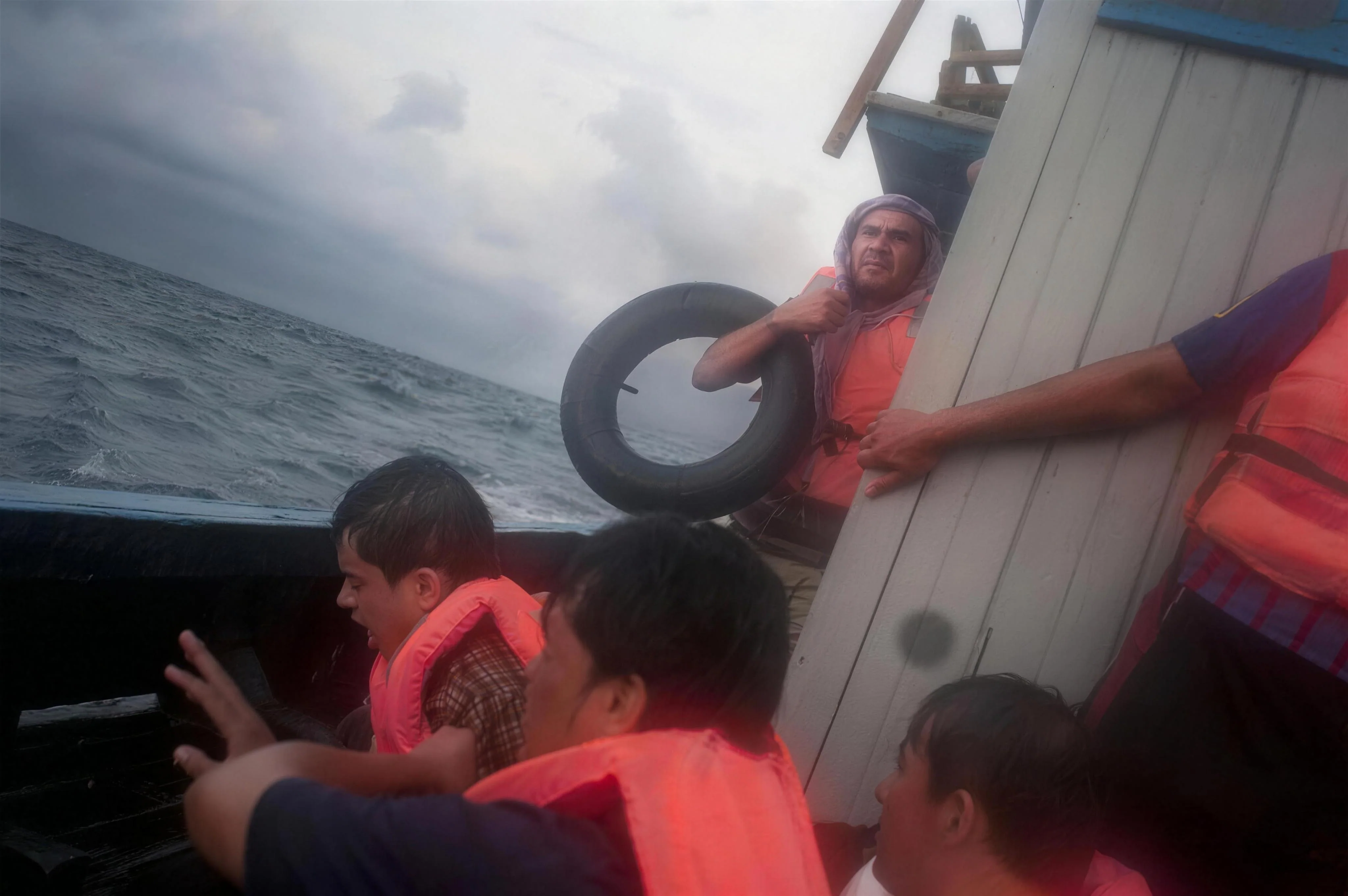 On the deck in heavy sea, a man holding a tyre tube and others in life jackets. From Seeking Asylum, 2012.