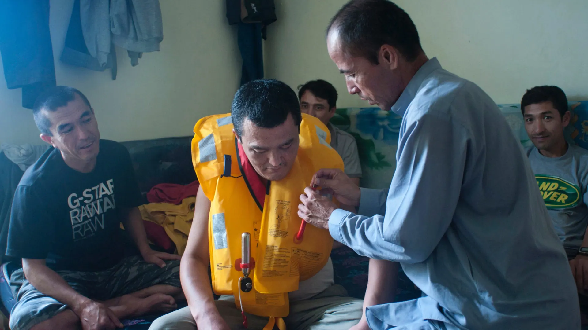 A man tries on a yellow life jacket in a crowded room while another adjusts the straps, Indonesia, 2012.